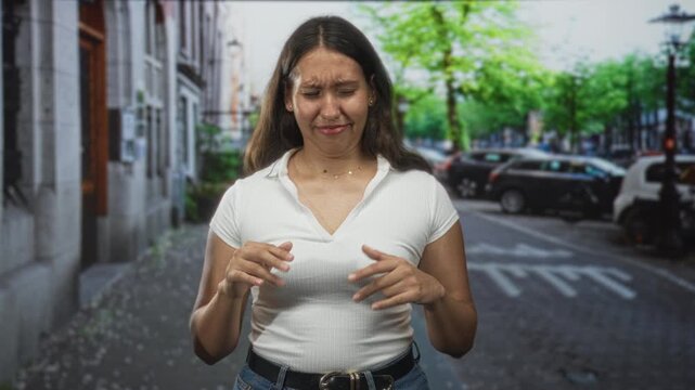 Woman gesturing with hands on a city street wearing a white ribbed top and blue jeans near parked cars and leafy trees; awkward conversation embarrassment.