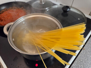 A bundle of dry spaghetti is being lowered into a stainless steel pot of vigorously boiling water on a black stovetop, next to a pan containing simmering tomato sauce.