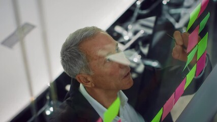 An elderly man with gray hair is actively writing on colorful sticky notes in a contemporary office environment. He displays focus and determination as he works on a business project. - Powered by Adobe
