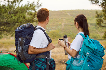 Couple hikers with backpacks near tent at sunset. They face the hills from a forest campsite,...