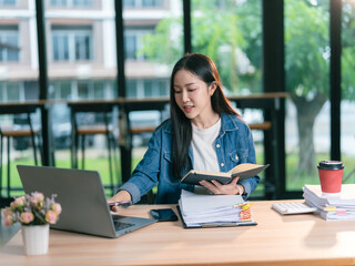 Fototapeta premium Asian woman working with laptop in office, holding pen and reading financial document, smiling and working at desk in office with computer.