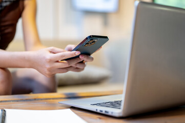 Close up of a woman's hands holding phone over laptop sitting at wooden table in living room or cafe