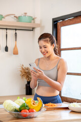Asian woman in sportswear holding phone standing beside kitchen cooking counter with fresh vegetable