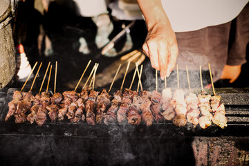 Street food seller grilling tasteful satay on skewers over hot and sizzling smokey charcoal. Authentic Asian culinary concept.