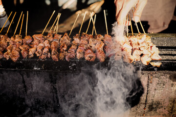 Street food seller grilling tasteful satay on skewers over hot and sizzling smokey charcoal. Authentic Asian culinary concept.