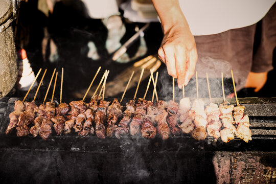 Street food seller grilling tasteful satay on skewers over hot and sizzling smokey charcoal. Authentic Asian culinary concept.