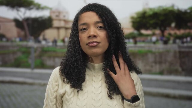 Woman touches curly hair wearing cable knit sweater by roman ruins with neutral expression; serenity.