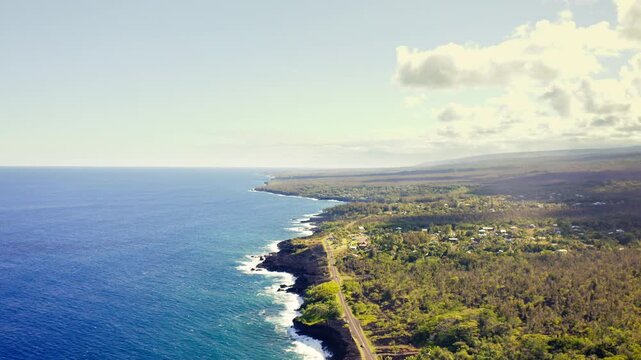 A narrow coastal road winds beside the deep blue Pacific, tracing the rugged volcanic shore of Hawaii&rsquo;s Big Island where waves meet green tropical forest under bright morning light.