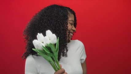 Young woman holds white tulips with hand and brightly smiles against bold red background in studio; hope.