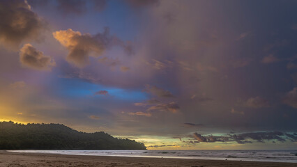 Beautiful evening landscape after sunset. Ocean waves are foaming on a sandy beach. The sky is illuminated with golden at the horizon. Purple and pink clouds. A hill in the distance. Malaysia.