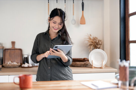 A smiling Asian woman happily using a digital tablet while standing in her contemporary kitchen.