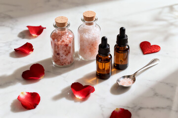 Small glass bottles of essential oils and corked jars of pink bath salts are arranged on a marble surface, surrounded by red rose petals and a glitter heart for a spa or relaxation concept.