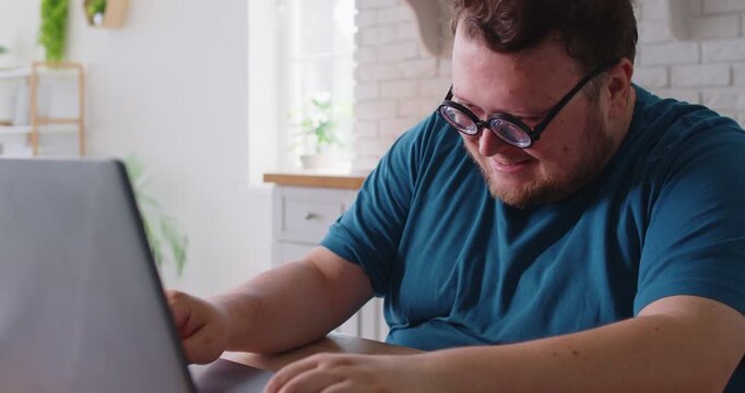 Obese funny man with creative idea typing online on laptop, sitting at home kitchen table. Happy chubby busy guy with round glasses using computer for social media chat and comment, looking at screen