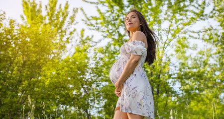 Pregnant woman holding showing belly baby bump smiling happy in 3rd trimester in spring forest nature. Asian expectant mom holding tummy.
