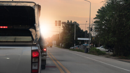 Rear side of transport pickup car silver color with turn on brake light. All cars are stopped at the traffic light ahead at the intersection. Light shining against the evening sky.