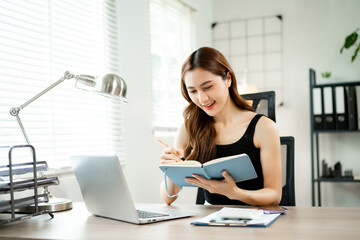 Smiling Asian woman working from home on laptop, writing notes, online learning or freelance business concept in home office.
