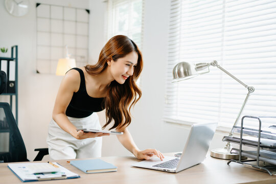 Smiling Asian woman standing in bright modern home office using smartphone, concept of digital lifestyle, communication, and remote working success.