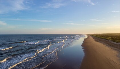 Ocean waves meet sandy beach under a soft blue sky