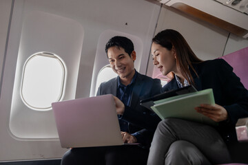 Asian business professionals working together on a laptop while sitting on a commercial airplane. They appear to be discussing a project or presentation in a productive and collaborative manner.