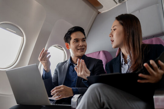 Asian businesspeople discussing work while flying on a plane. The man gestures as he explains something, while the woman listens holding a folder and pen, showing in-flight collaboration.
