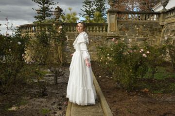 Fototapeta premium portrait of beautiful young blonde model wearing historical white wedding gown. ethereal back view figure, walking away in romantic rose garden of fairytale castle garden background