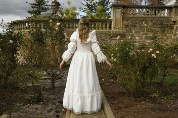 Fototapeta premium portrait of beautiful young blonde model wearing historical white wedding gown. ethereal back view figure, walking away in romantic rose garden of fairytale castle garden background