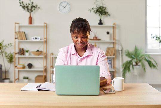 Portrait of young smiling African American woman sitting at desk, using laptop computer for work or studying. Female college student, businesswoman or office worker learning or working online.