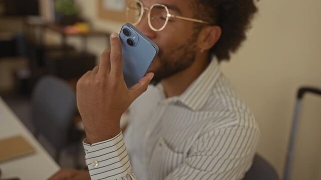 Man speaking into smartphone in modern office wearing glasses and striped shirt highlighting technology usage among professionals in indoor workplace setting with focus on communication.
