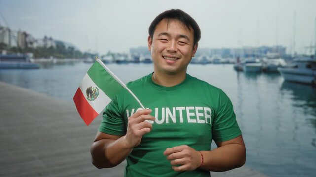 Young man holding mexican flag by the seaside, wearing a green volunteer shirt, showcasing a cheerful expression outdoors at the port.