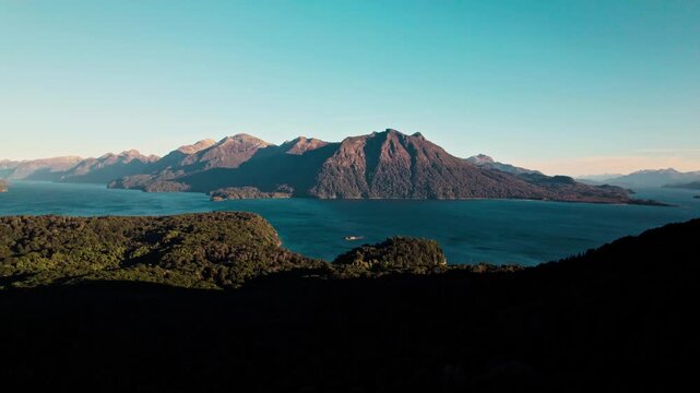Epic wide shot of the vast glacial lake (Lago Nahuel Huapi region) surrounded by dark, forested mountain islands under a clear blue sky.