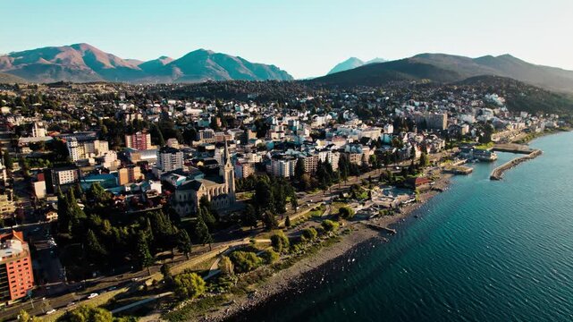 High-angle drone shot capturing the urban core of San Carlos de Bariloche, Argentina, with the historic Cathedral nestled by the vast Nahuel Huapi Lake and the dramatic Patagonian Andes mountains.
