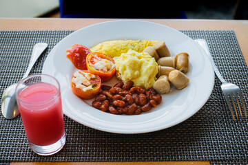 Hotel healthy breakfast menu of mashed potatoes, omelet, read beans, tomatoes, and sausages served on a white plate with a glass of guava juice