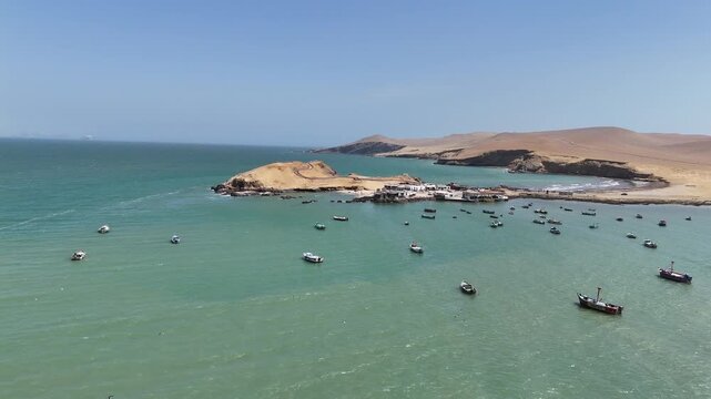 Aerial moving forward captures a high-angle view of the small fishing village at Playa Lagunillas cove in Peru's Paracas National Reserve, with boats anchored in the bay