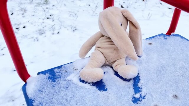 Lonely rabbit doll sitting alone on carousel at winter. Sad forgotten abandoned rabbit sitting outside in snowy day, lost toy waiting for owner. Loneliness concept, International missing Children day