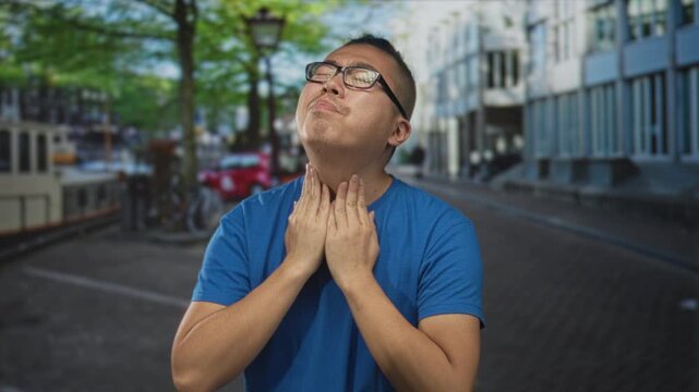 Young chinese man in blue t shirt clutching neck and grimacing on a tree lined city street near canal and parked bicycles; discomfort.