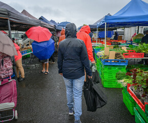 Couple walking in the farmer’s market in the rain. Unrecognizable people with umbrellas shopping in the market. Auckland.