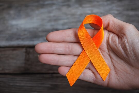 Hand holding an orange awareness ribbon against a wooden background, representing support for various health conditions and causes