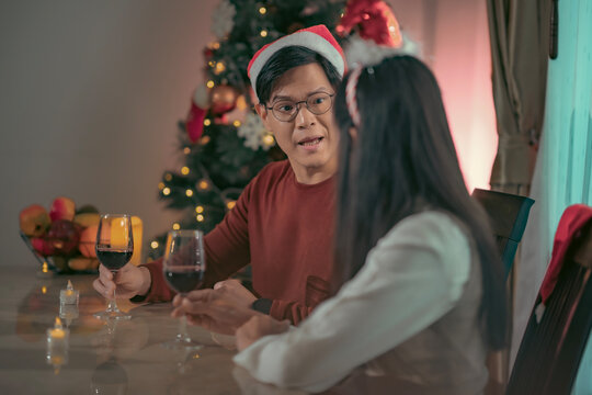 A warm and romantic Christmas moment featuring an Asian couple celebrating together at home. Asian couple wearing Santa hats holding wine glasses and talking during cozy Christmas celebration at home.