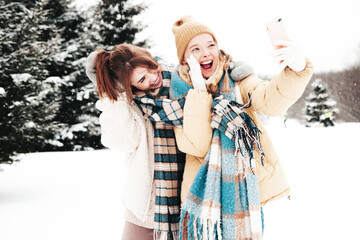 Two young beautiful smiling hipster female in trendy warm clothes and scarfs.Carefree women posing in the street in park. Positive pure models having fun in snow. Enjoying winter moments.Taking selfie