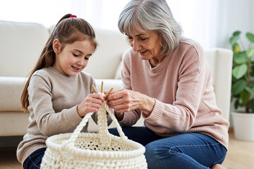A caring grandmother patiently teaching her happy young granddaughter how to knit or crochet with white yarn in a cozy living room