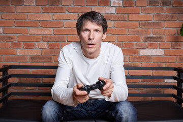 A middle-aged man gamer holds a gamepad and plays an online game on a computer.