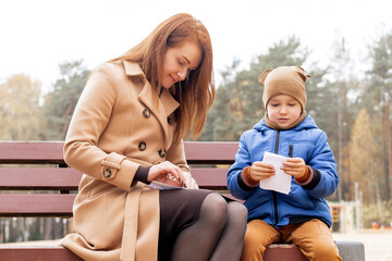 Mother and Son Folding Paper Boat Together on Park Bench During Autumn Day