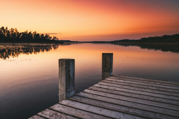 Obraz premium A wooden pier juts into a calm lake at sunrise or sunset, reflecting the stunning gradient of orange, pink, and red across the water, framed by a dark forested horizon.