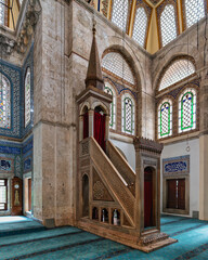 Interior of Muradiye Mosque, Manisa, Turkey, showcasing its ornate wooden minbar, blue patterned carpet, and vibrant stained glass windows with intricate Islamic calligraphy
