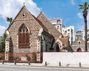 Bishop Collins Memorial Hall, St. John's Cathedral, Izmir, Turkey. Historic stone church with Gothic window, roof crosses, and palm trees on a sunny day