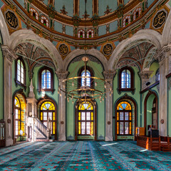 Ornate sunlit interior of Salepcioglu Mosque, Izmir, Turkey. Features a grand dome, Islamic calligraphy, large chandelier, vibrant stained glass windows, and a patterned turquoise carpet