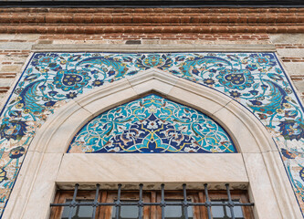 Exterior of Konak Mosque, Izmir, Turkey. Close-up of Iznik blue and turquoise floral tilework above a barred window, set within an arched frame and brick wall