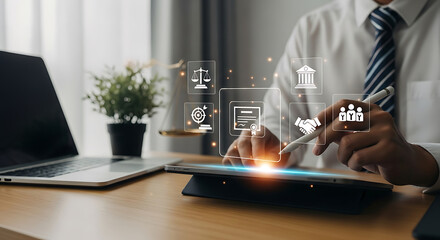 Man uses stylus on tablet displaying legal and business concept icons at desk with laptop and scales of justice