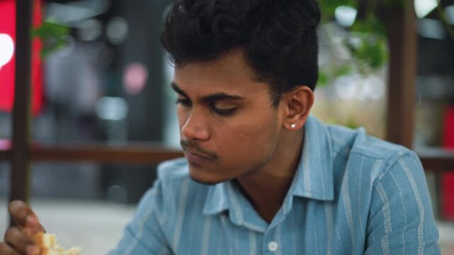Close up view of man with dark fluffy hair biting into juicy burger, lips and fingers visible, background blurred, natural light highlighting texture of bun and meat in candid eating shot, moody