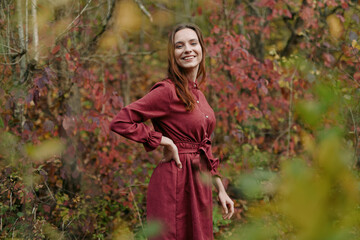 A smiling young woman in a simple red dress stands among autumn foliage, photographed with genuine...